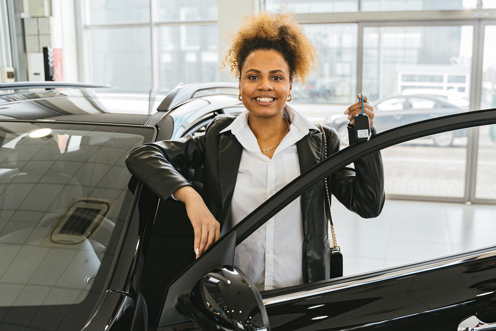 Smiling woman with car keys standing beside her new vehicle in a dealership showroom, auto